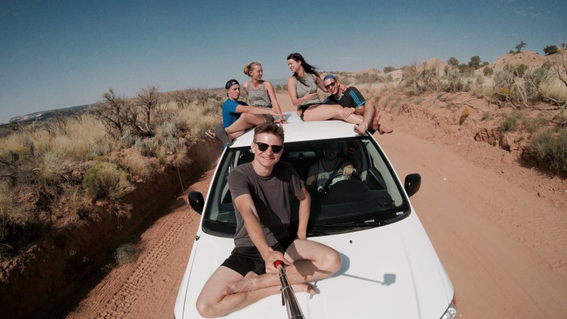 Work and travel – Jak si správně vybrat agenturu a jaký je rozdíl mezi zprostředkovateli. Parta mladých lidí na autě s akční kamerou na selfie tyči uprostřed pouště v USA.